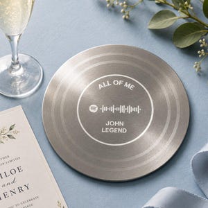 Könnte beinhalten: Ein silberfarbener Untersetzer mit dem Text "ALL OF ME" und "JOHN LEGEND", der einen Spotify-Code umgibt. Der Untersetzer hat ein plattenähnliches Design mit konzentrischen Rillen. Ein Sektglas und eine Hochzeitseinladung befinden sich im Hintergrund.
