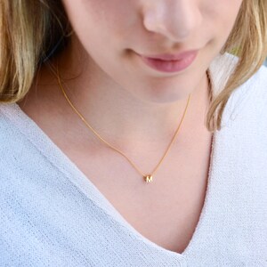May include: A delicate gold-colored necklace with a small pendant featuring the letter "M". The necklace is worn around the neck, resting on a white sweater. The chain is thin and the pendant is a simple, minimalist design.