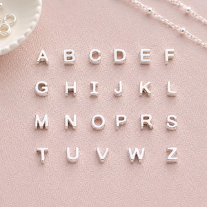 May include: Silver alphabet charms, capital letters A through Z, arranged on a pink surface. The letters are evenly spaced and appear to be made of metal. A silver necklace with small beads is visible in the background.