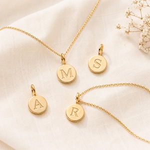 May include: Gold-colored necklaces with circular pendants, each displaying a different letter: M, S, A, and R. The pendants are suspended from delicate chains, set against a soft, off-white background. The jewelry is simple and elegant.