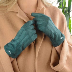 May include: Emerald green gloves with vertical stitching details. The gloves have the initials "AL" embroidered in white on each cuff. The gloves are made of a soft material and are worn by a person wearing a tan coat.