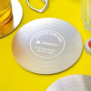 Op de afbeelding: Een zilverkleurige ronde onderzetter met de tekst "CUSTOM SONG HERE" en "CUSTOM BAND/ARTIST HERE" in een cirkelvormig ontwerp. De onderzetter heeft een Spotify-code in het midden. Andere onderzetters, een flesopener en een glas zijn zichtbaar.