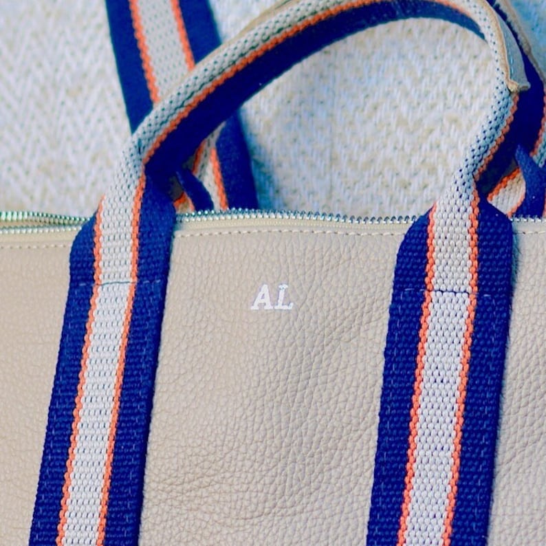 May include: Beige leather tote bag with blue and white striped straps. The bag has the initials "AL" embossed on the front.