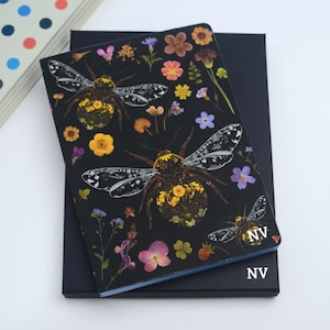 Può includere: Un taccuino nero con un design floreale e di insetti. La copertina presenta un'illustrazione dettagliata di un'ape con dettagli floreali e vari fiori colorati. Il taccuino è sopra una scatola blu scuro, con le iniziali "NV" nell'angolo.
