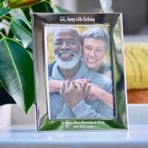 Puede incluir: Marco de fotos plateado con una inscripción personalizada que dice "Gill, Happy 60th Birthday" y "So Many More Memories to Make, Love Nick xxxx". El marco contiene una foto de una pareja mayor sonriendo y abrazándose.