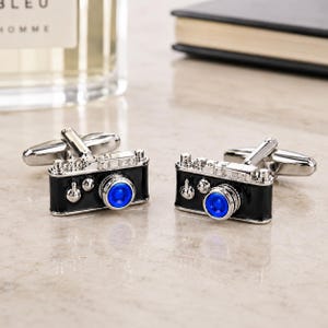 May include: A pair of silver-toned cufflinks shaped like vintage cameras. Each cufflink features a glossy black body, a prominent circular blue lens, and silver metallic detailing on the top and sides, complete with a standard swivel toggle back.