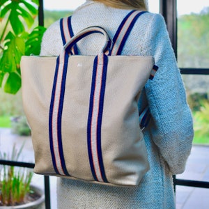 A beige leather backpack with navy blue and orange striped straps. The backpack has a zipper closure and a top handle. The initials "AL" are monogrammed on the front of the bag.