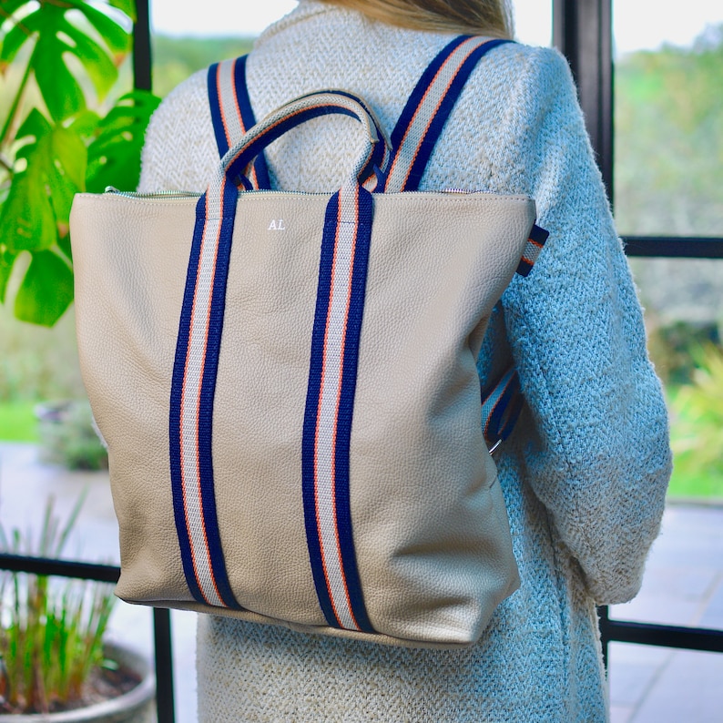 A beige leather backpack with navy blue and orange striped straps. The backpack has a zipper closure and a top handle. The initials "AL" are monogrammed on the front of the bag.