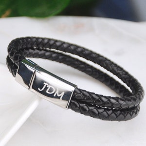Pode incluir: Um bracelete de couro preto com um fecho prateado gravado com as iniciais "JDM".