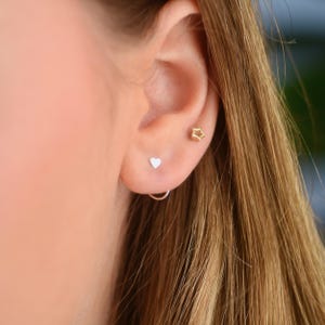 Puede incluir: Primer plano de la oreja de una persona con tres pequeños pendientes. Los pendientes son una estrella dorada, un corazón plateado y un aro plateado.