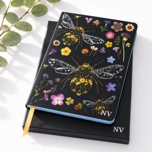 Peut inclure: Journal noir avec un motif d'abeille floral et un signet en ruban doré. Le corps de l'abeille est rempli de fleurs jaunes et les ailes sont blanches avec un motif délicat. Le journal est sur une boîte noire avec les initiales "NV".