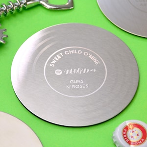 May include: A silver-colored metal coaster with the text "Sweet Child O' Mine" and "Guns N' Roses" engraved on it. The coaster also features a Spotify logo and a soundwave graphic. The coaster is round and has a brushed metal finish.