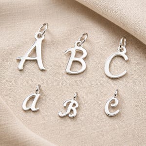 May include: Silver alphabet charms, including uppercase A, B, and C, and lowercase a, b, and c. Each letter has a small loop at the top for attaching to a necklace or bracelet. The charms are displayed on a neutral-colored fabric.