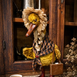 Puede incluir: Una figura de pato de peluche amarillo con un gorro beige de volantes, gafas redondas de montura metálica y una bufanda de cuadros marrones y azul marino sujeta con un broche ovalado rojo. El pato lleva botas rojo oscuro y un bolso.