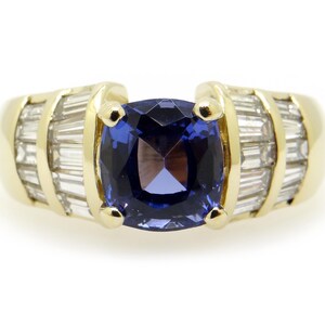 May include: A gold ring featuring a large, square, deep blue gemstone as the centerpiece. The ring is flanked by rows of smaller, rectangular, clear gemstones. The setting is a polished gold.