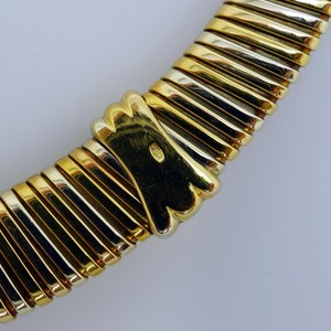 May include: A gold and silver necklace with a textured, ribbed design. The necklace is made of two different metals, one gold and one silver. The necklace is curved and has a clasp at the end.