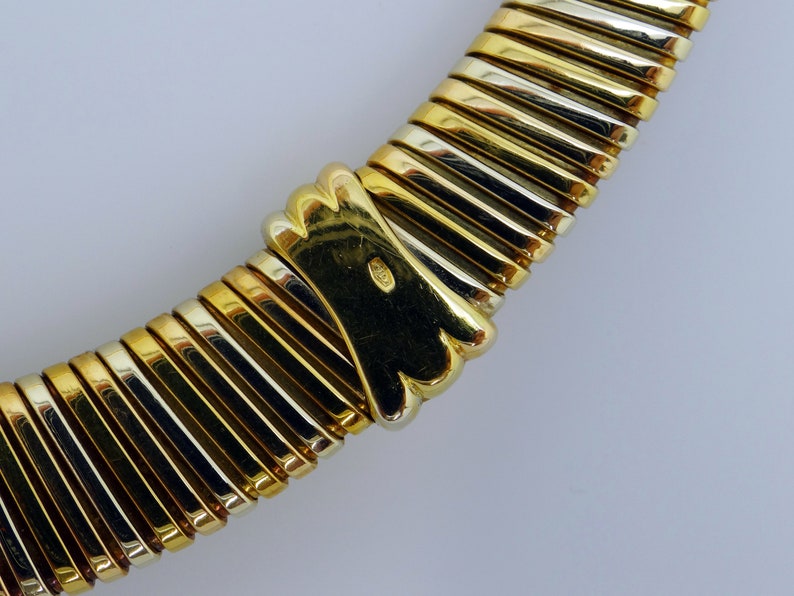 May include: A gold and silver necklace with a textured, ribbed design. The necklace is made of two different metals, one gold and one silver. The necklace is curved and has a clasp at the end.