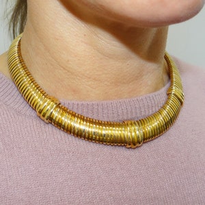 May include: A gold-toned necklace with a thick, rigid design. The necklace is made of multiple linked rings that create a smooth, rounded shape.