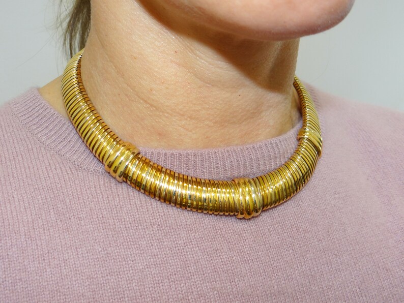 May include: A gold-toned necklace with a thick, rigid design. The necklace is made of multiple linked rings that create a smooth, rounded shape.