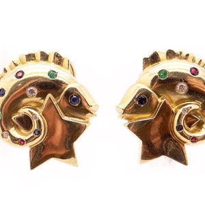 May include: A pair of gold earrings shaped like a ram's head with a star. The earrings are adorned with colorful gemstones, including rubies, emeralds, sapphires, and diamonds.