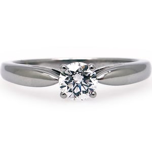 May include: A solitaire engagement ring with a round-cut diamond set in a polished, silver-colored band. The band has a smooth, rounded design that tapers towards the diamond. The diamond is held in place by four prongs.