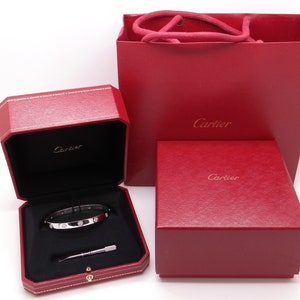 May include: A Cartier bracelet in a red box with a red Cartier shopping bag. The bracelet is silver with small diamonds.