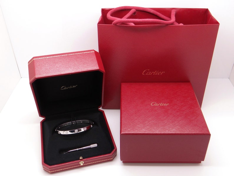 May include: A Cartier bracelet in a red box with a red Cartier shopping bag. The bracelet is silver with small diamonds.