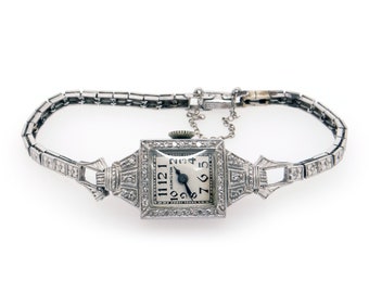 May include: A vintage Hamilton wristwatch with a square face and diamond-encrusted bezel. The watch has a white face with black Roman numeral hour markers and black hands. The watch is on a silver bracelet with diamond accents.