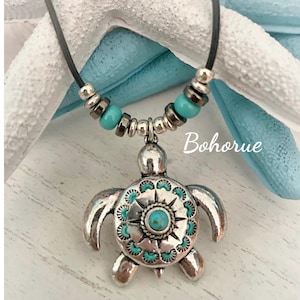 May include: A turquoise and silver turtle pendant necklace with a black leather cord. The turtle has a turquoise stone in the center of its shell and a silver starburst design.