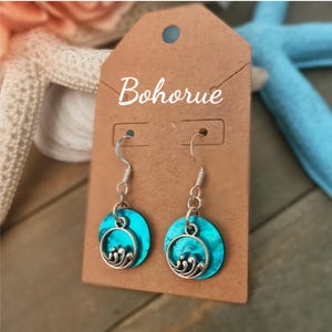 Op de afbeelding: Een paar turquoise blauwe oorbellen met zilveren golfontwerpen die aan zilveren haken hangen. De oorbellen worden getoond op een bruine kaart met het woord "Bohorue" erop gedrukt.