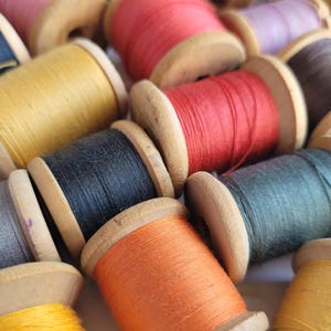 May include: A collection of wooden spools wrapped in various colors of sewing thread, including red, orange, yellow, black, grey, dark green, and brown. The spools are arranged in a cluster, showing the textured thread wound around the wooden cores.