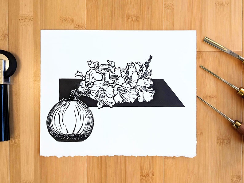 Bold black and white original linoleum block print of floral still life in a small round vase on pale wood background with carving tools.