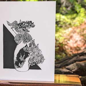 Bold black and white original linoleum block print of floral still life bouquet in a decorative vase sits on a small wood table outside amidst a peaceful redwood forest.