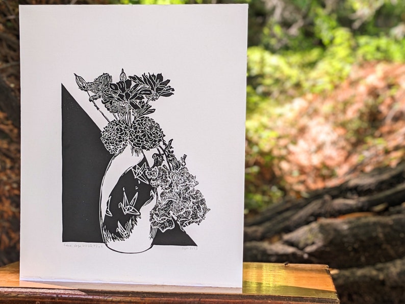 Bold black and white original linoleum block print of floral still life bouquet in a decorative vase sits on a small wood table outside amidst a peaceful redwood forest.