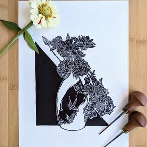 Bold black and white original linoleum block print of floral still life bouquet in a patterned vase. Print lies on pale wood background and includes two carving tools and a zinnia flower to show scale.