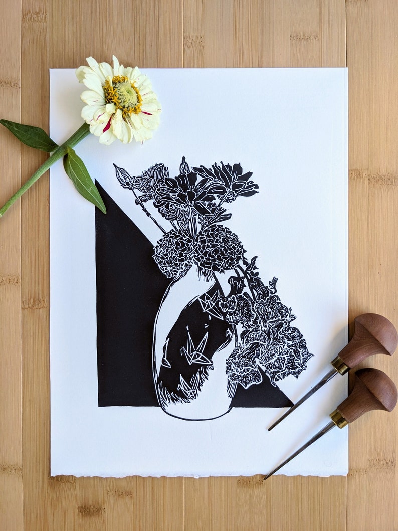 Bold black and white original linoleum block print of floral still life bouquet in a patterned vase. Print lies on pale wood background and includes two carving tools and a zinnia flower to show scale.