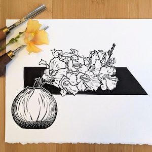 Bold black and white original linoleum block print of floral still life in a small round vase on pale wood background with carving tools and a Chantilly Light Snapdragon flower.