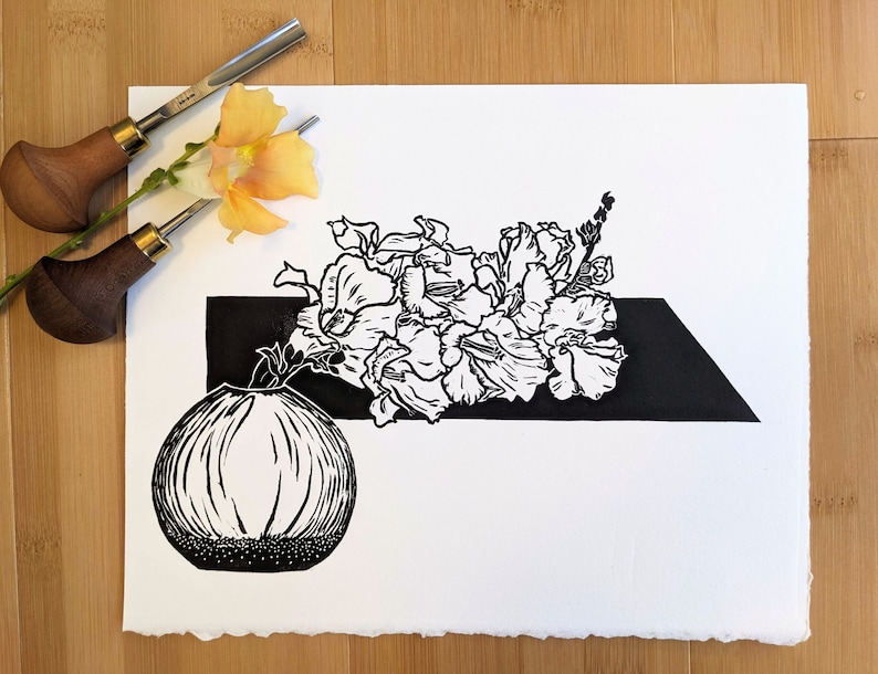 Bold black and white original linoleum block print of floral still life in a small round vase on pale wood background with carving tools and a Chantilly Light Snapdragon flower.