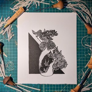 Bold black and white original linoleum block print of floral still life bouquet in a patterned vase on work desk surrounded by the tools which helped make it.