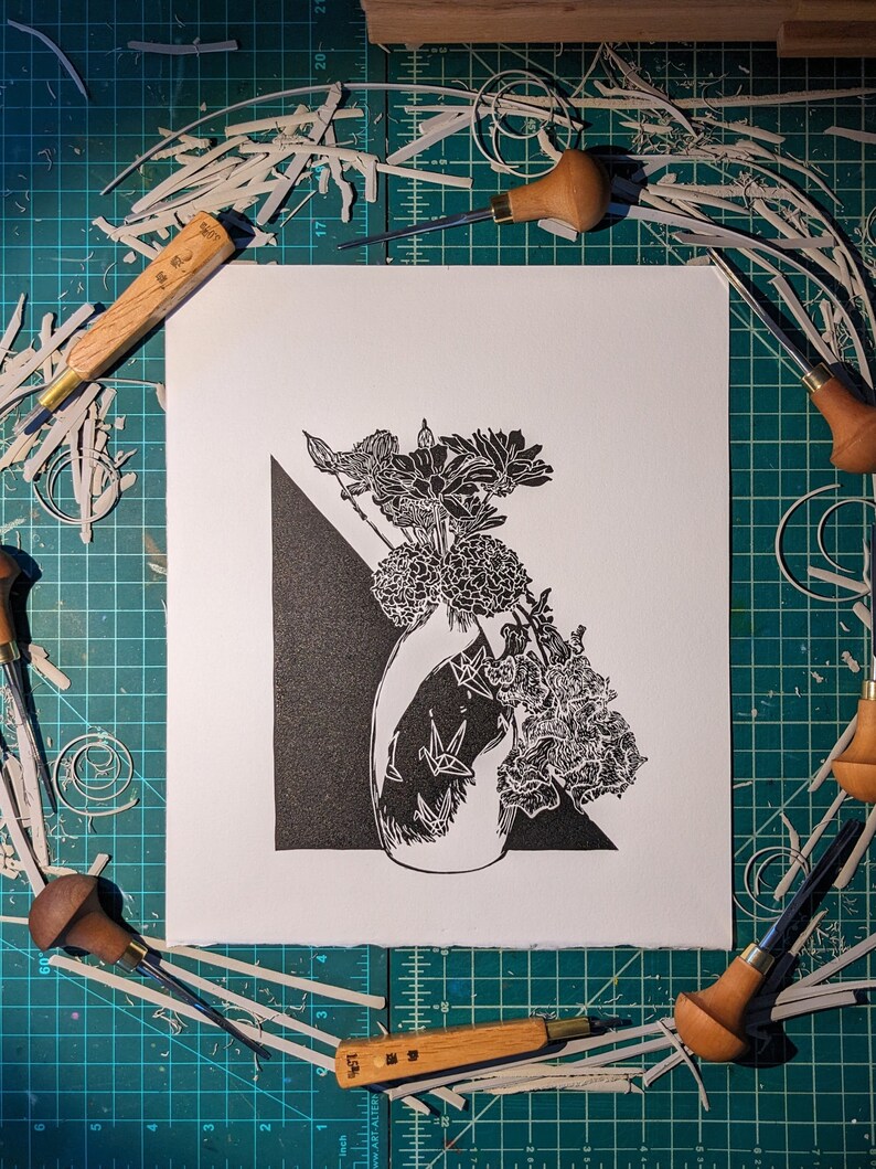 Bold black and white original linoleum block print of floral still life bouquet in a patterned vase on work desk surrounded by the tools which helped make it.