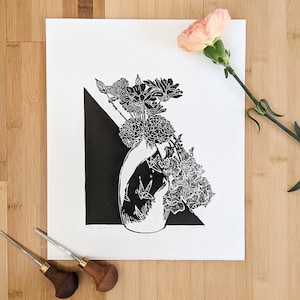 Bold black and white original linoleum block print of floral still life bouquet in a decorative vase. Print lies on a wooden surface beside a carnation and carving tools.