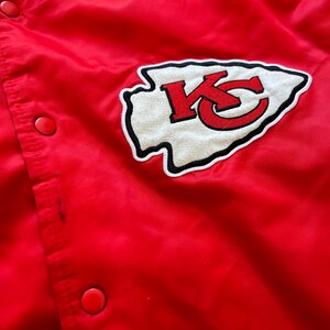 May include: Red satin bomber jacket with a white and red embroidered Kansas City Chiefs logo on the front. The jacket has a ribbed collar and cuffs.