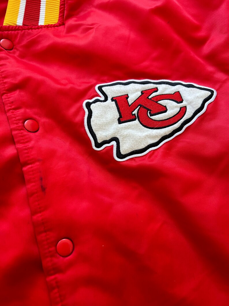 May include: Red satin bomber jacket with a white and red embroidered Kansas City Chiefs logo on the front. The jacket has a ribbed collar and cuffs.