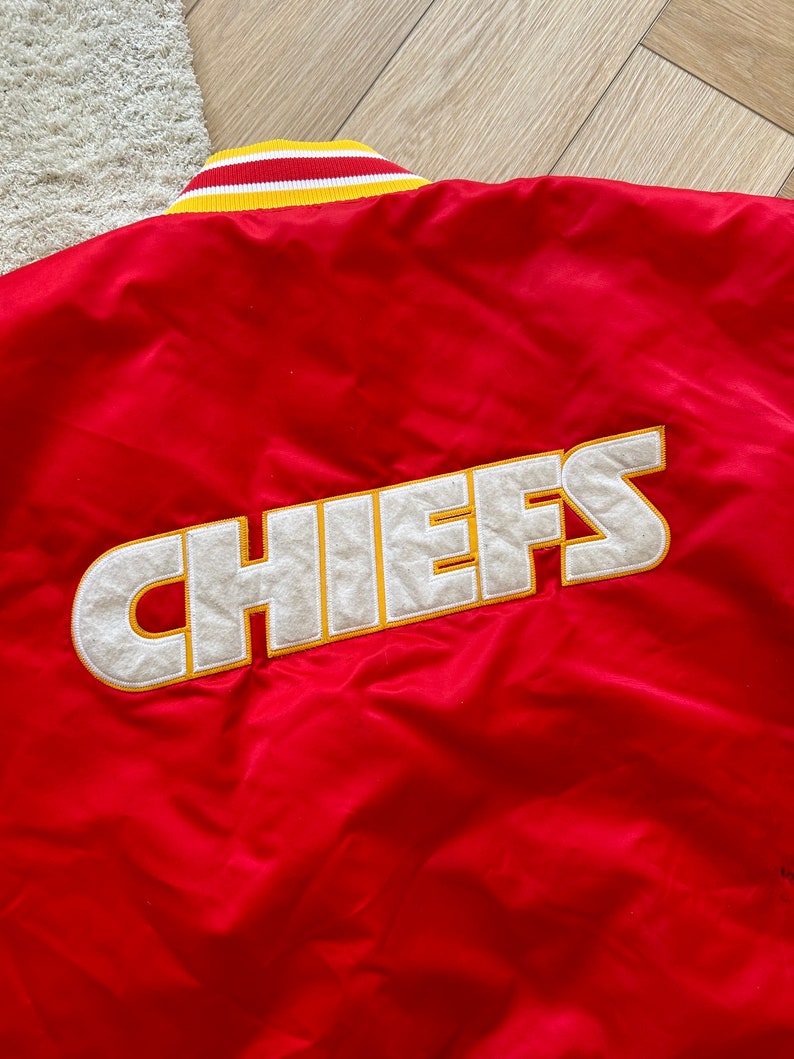 May include: Red satin bomber jacket with a white and yellow striped collar and the word "CHIEFS" embroidered in white with a yellow outline on the back.