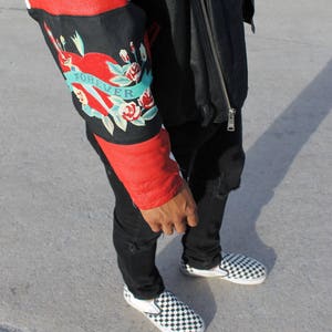 May include: A red and black leather jacket with a floral design on the sleeve. The design includes a heart with the word "FOREVER" written on it. The jacket is worn over a black shirt and black jeans. The person is wearing black and white checkered slip-on shoes.