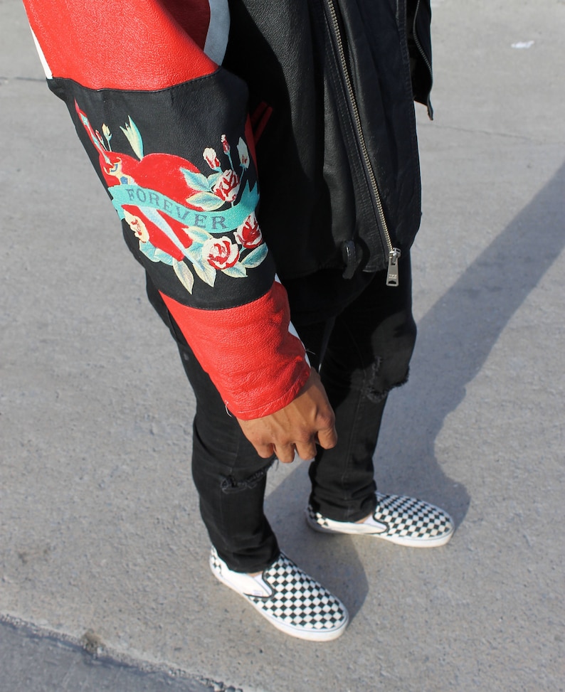 May include: A red and black leather jacket with a floral design on the sleeve. The design includes a heart with the word "FOREVER" written on it. The jacket is worn over a black shirt and black jeans. The person is wearing black and white checkered slip-on shoes.