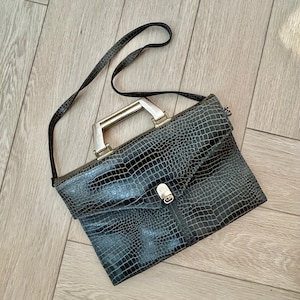 May include: A dark gray, faux crocodile-skin briefcase with a silver-toned handle and clasp. The bag has a long, thin shoulder strap. The briefcase is rectangular and has a textured surface.