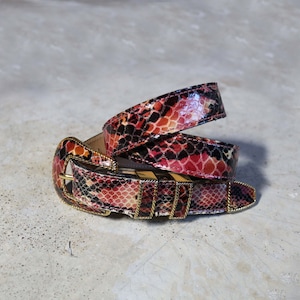 Peut inclure: Une ceinture en cuir à motif serpent rouge, noir et blanc avec une boucle dorée.
