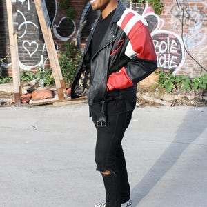 May include: A man wearing a black leather jacket with red and white accents, black jeans with a distressed knee, and black and white checkered slip-on shoes.
