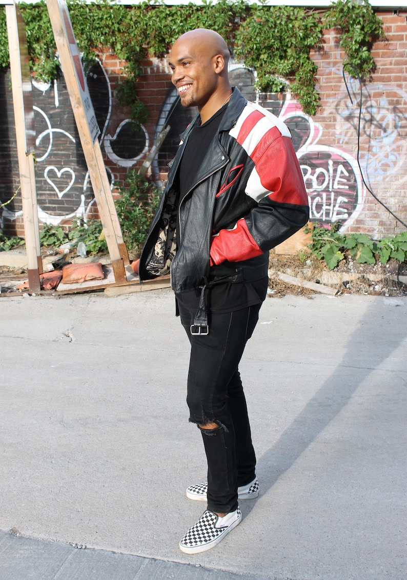 May include: A man wearing a black leather jacket with red and white accents, black jeans with a distressed knee, and black and white checkered slip-on shoes.